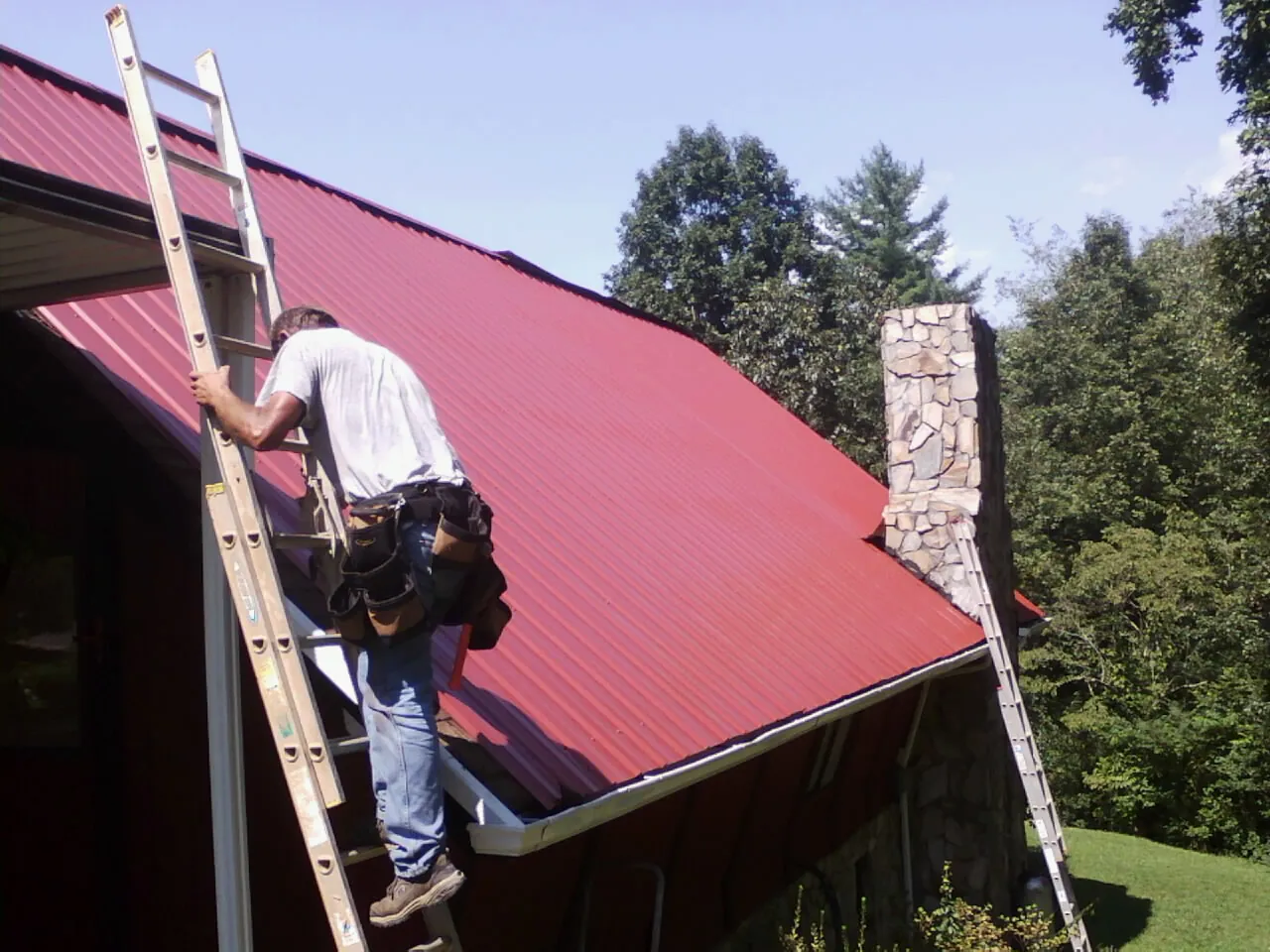 Roofer on a ladder beside a red metal roof and stone chimney — Soffit & Fascia Repair by Roofing Brothers in Ford